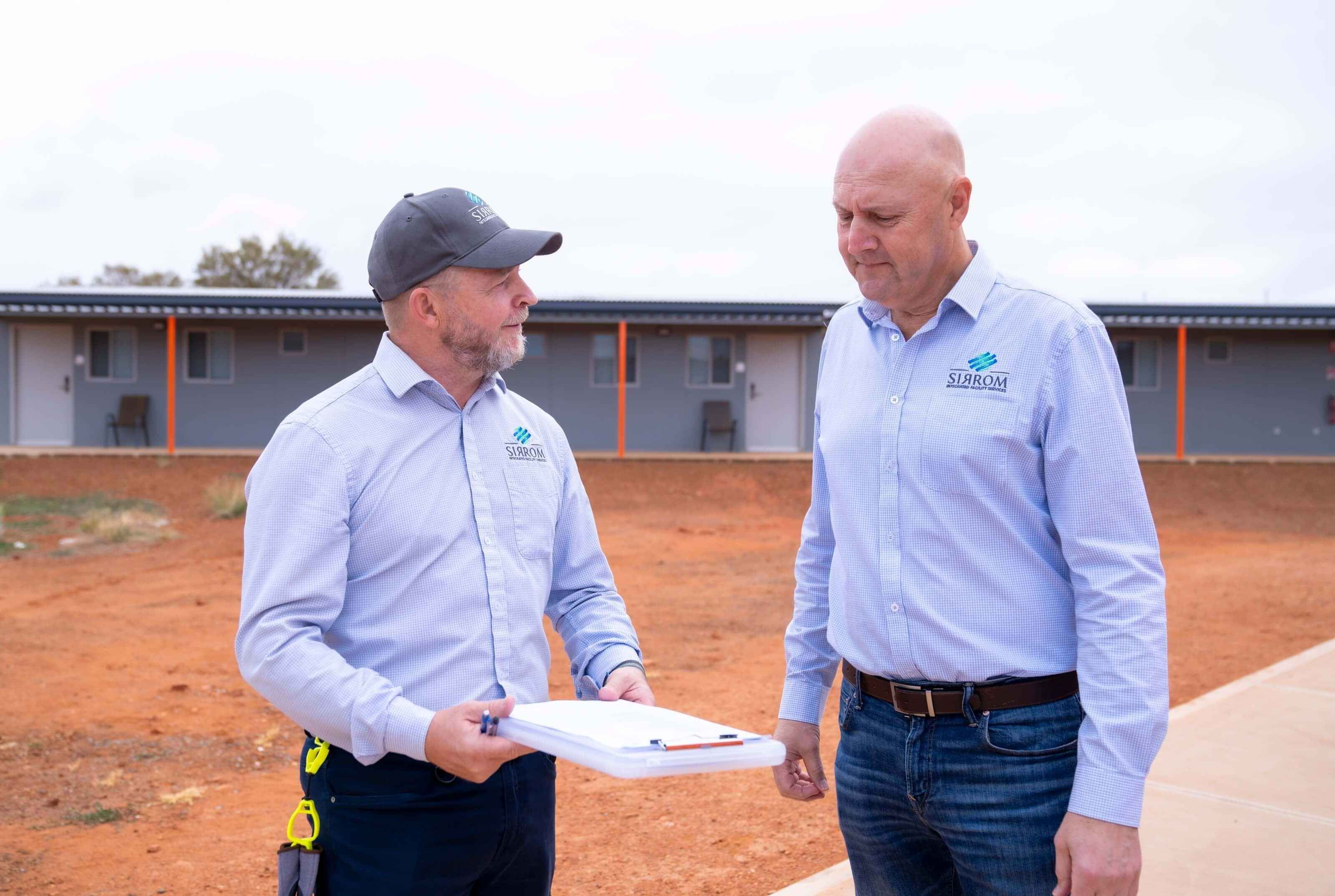 One man is holding a clipboard while talking to another man in front of a building in the desert.