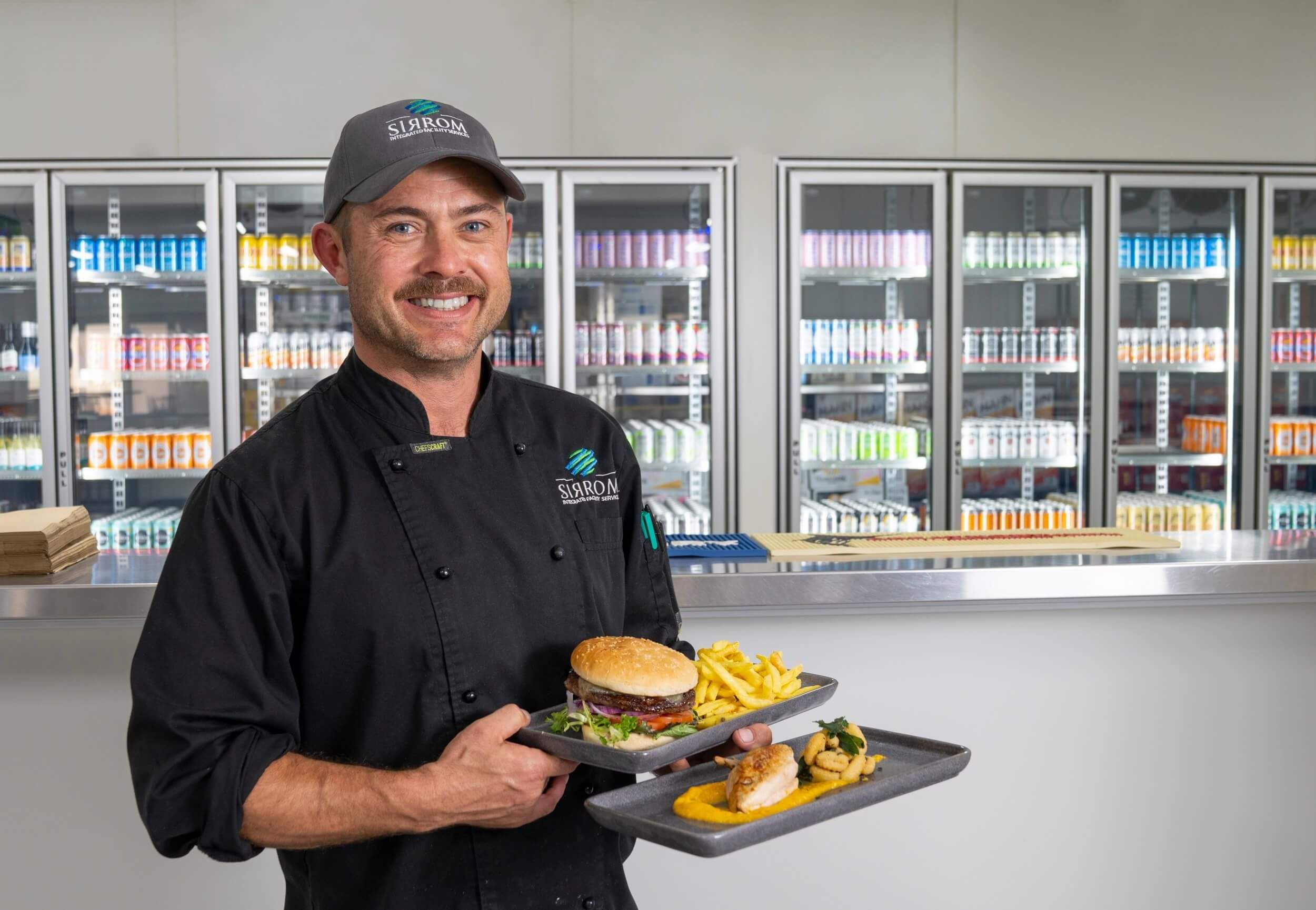 a male chef is holding two plates of well-presented food in front of a bar, with alcoholic drinks behind.
