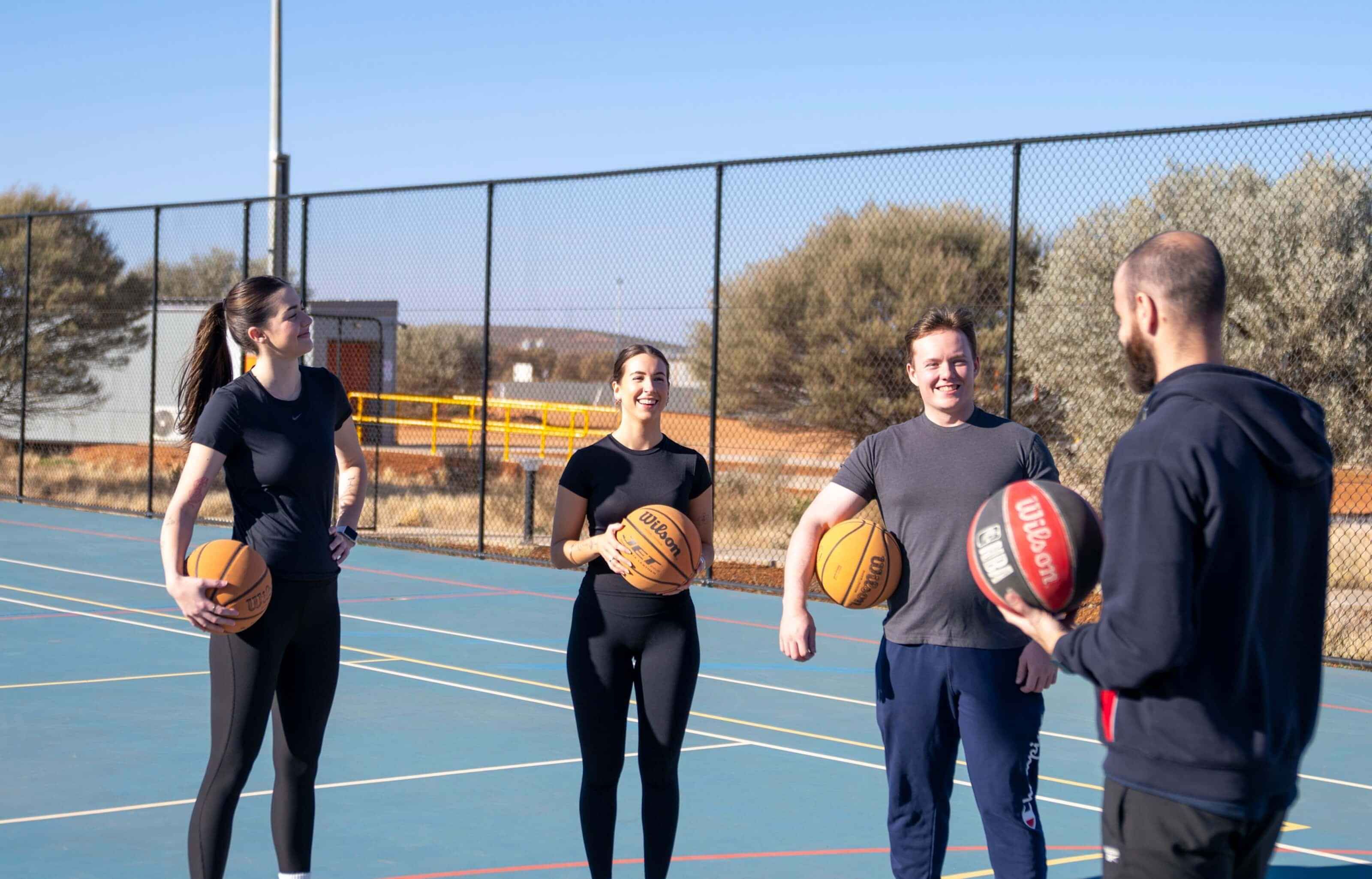 A group of people wearing gym clothes standing together hold basketballs and are smiling.