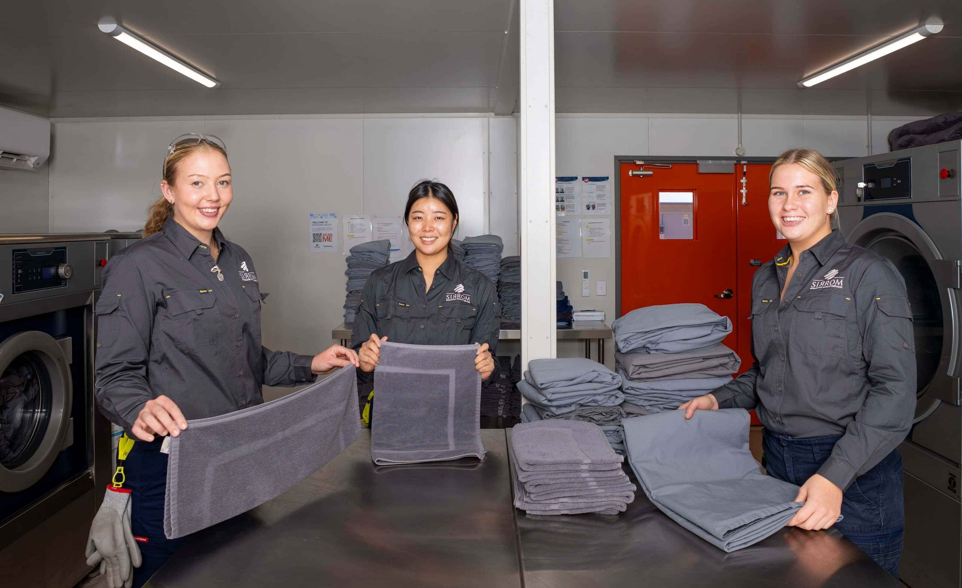 A group of smiling young women neatly fold towels in a commercial laundry.