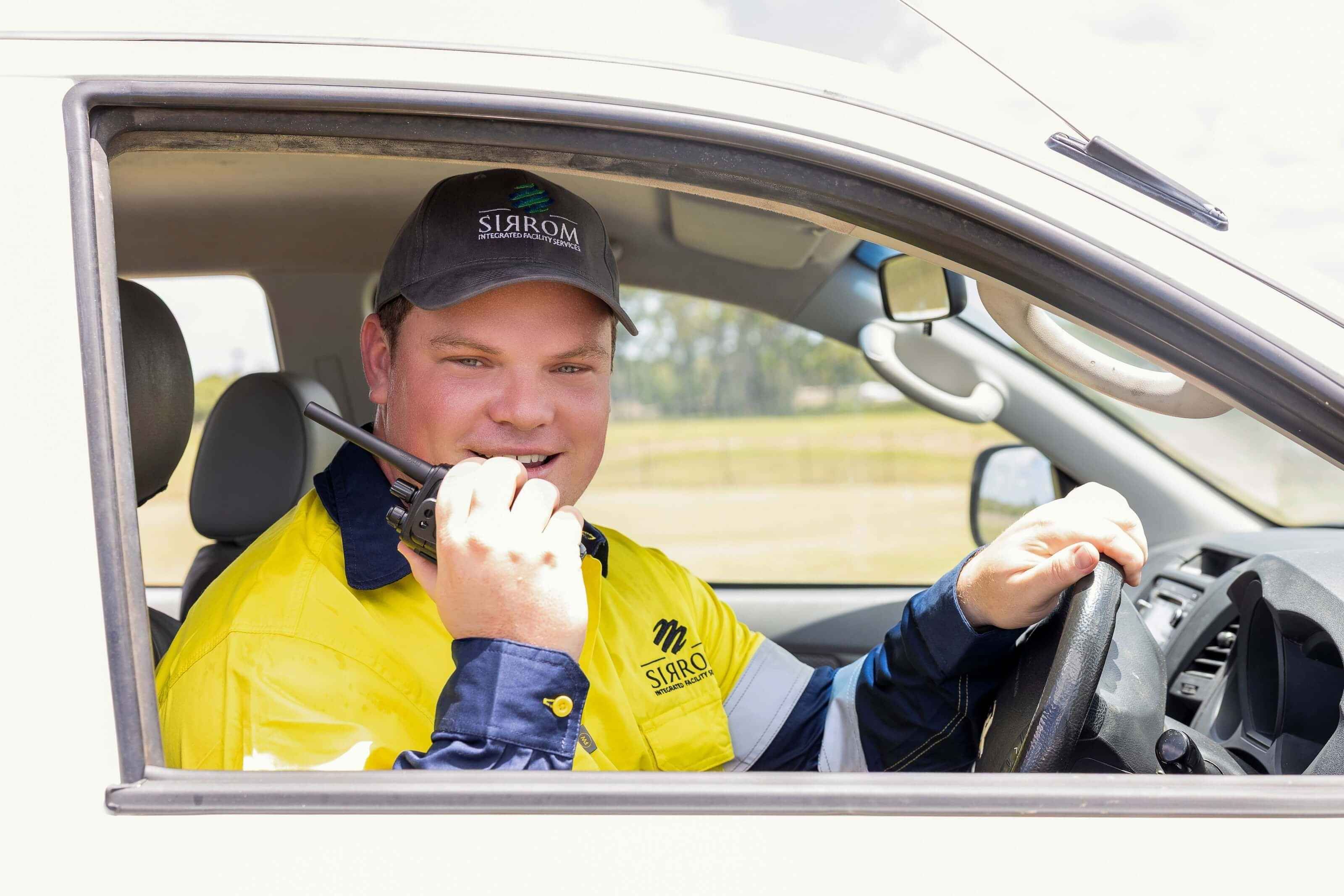 A man wearing PPE talks into a walkie talkie while driving a car.