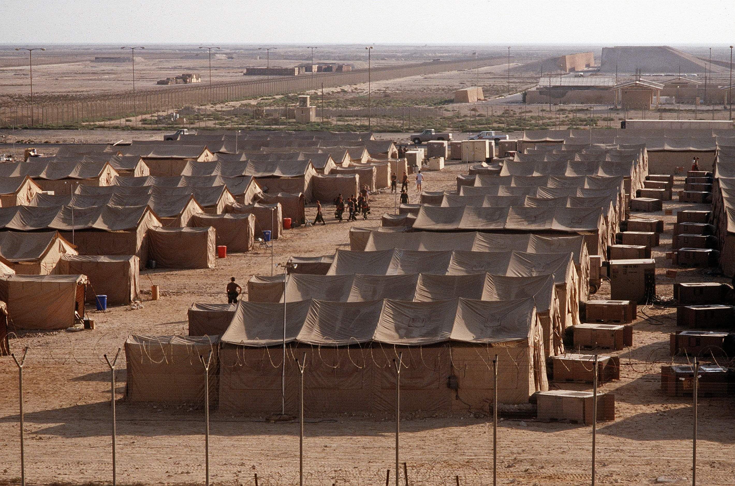 A camp of army tents surrounded by security fences in a desert environment.
