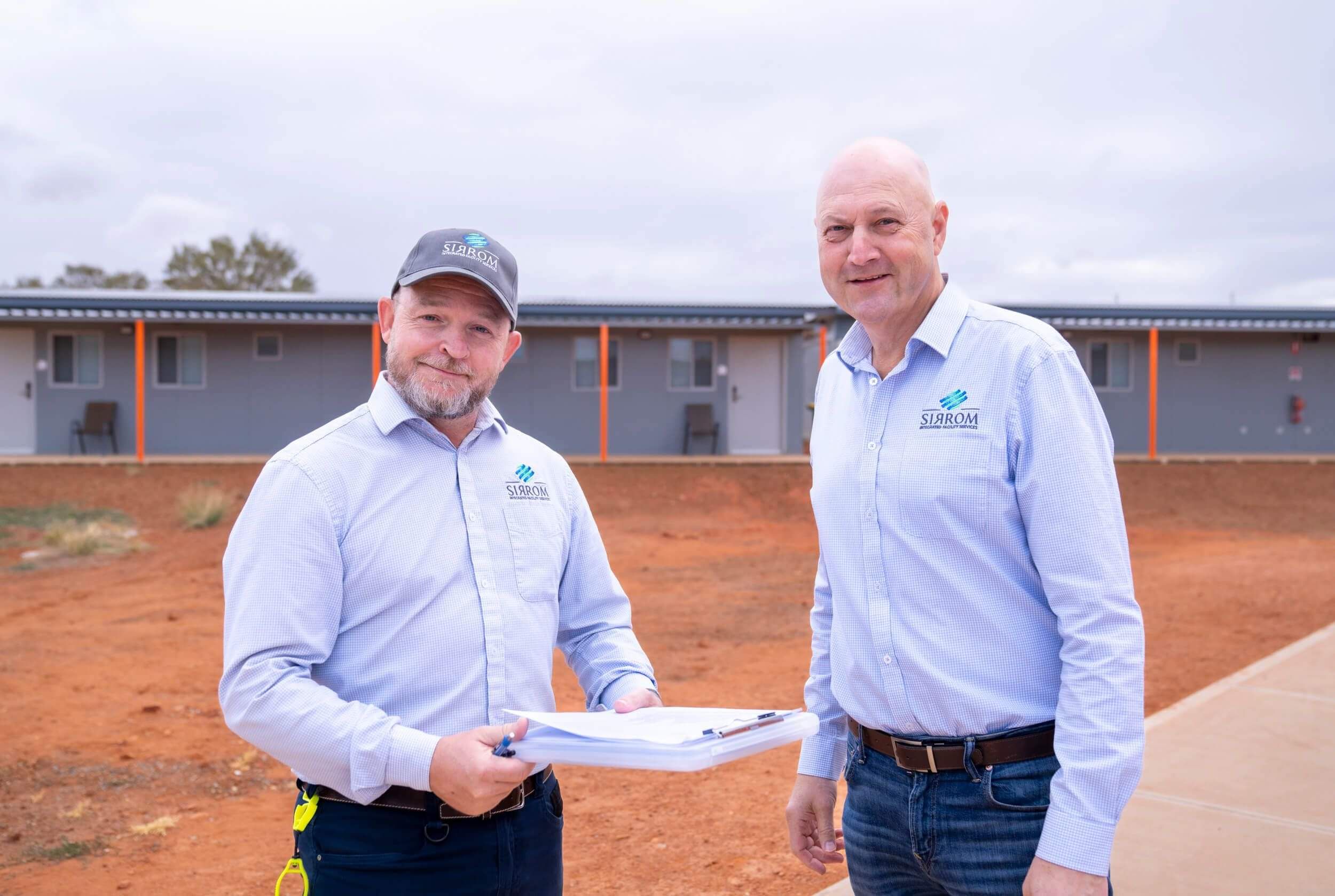 Two men are standing together in front of a group of buildings, with one man holding a clipboard.