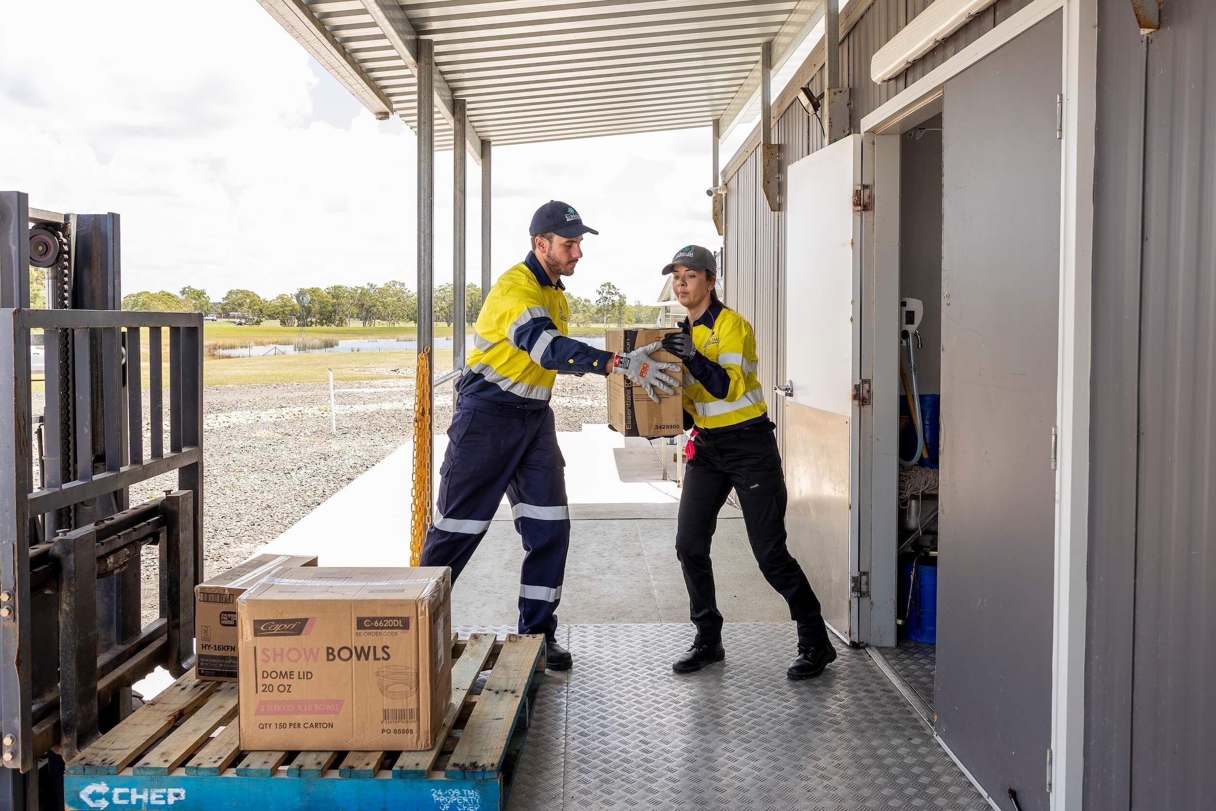 Wearing PPE, a man passes a woman a cardboard box in a loading dock.
